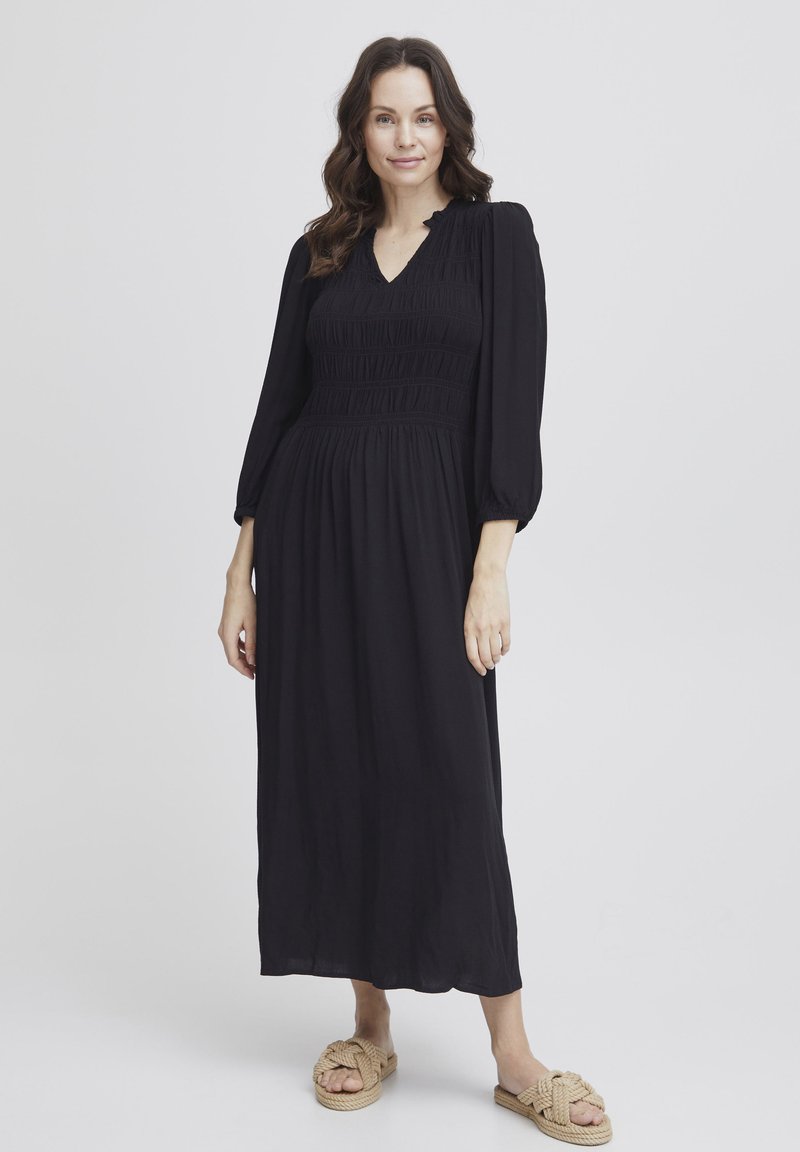 Schwarzes, langärmeliges Kleid mit V-Ausschnitt, gerafftem Oberteil und fließendem Rock. Getragen mit hellbraunen, geflochtenen Schlupfsandalen.