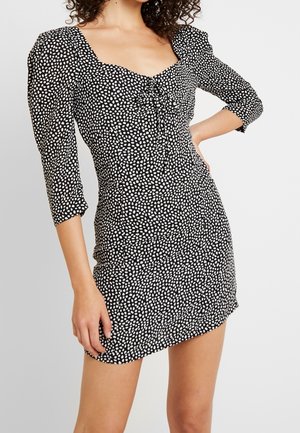 Petite robe noire à pois blancs irréguliers, devant froncé, épaules bouffantes et manches trois-quarts sur une femme debout.