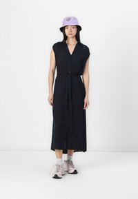 Abito camicia nero con cintura in vita, senza maniche e lunghezza midi, abbinato a un cappello a secchiello lilla e sneakers grigie.