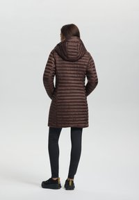 Brauner Puffer-Mantel mit Kapuze, ausgestattet mit einer gesteppten Textur und vertikalen Streifen, kombiniert mit schwarzen Leggings und schwarzen Schuhen.