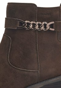 Bottine en daim marron avec un motif décoratif en chaîne métallique sur le dessus. Elle présente des coutures et une semelle texturée pour une meilleure adhérence.