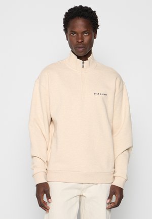 Mand iført en beige sweatshirt med kvart-zip og "LYLE & SCOTT" logo på brystet, kombineret med lyse bukser, står foran en ensfarvet baggrund.