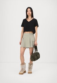 Melna cropped tops, gaiši zaļa plisēta svārki ar sānu kabatu, brūnas zābaku ar pūku apdari, un zaļa auduma rokassoma ar divām priekšējām kabatām.