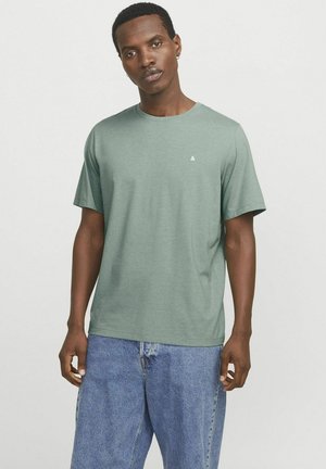 T-shirt vert à manches courtes avec col rond, fabriqué en tissu doux. Présente un petit logo sur la poitrine et une coupe décontractée. Porté avec un jean bleu.