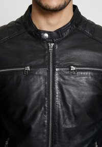 Zwarte leren jas met een hoge kraag, zilveren ritsen en twee ritszakken op de borst. Bevat getextureerde schouderaccenten en een gladde afwerking.