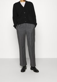 Černý pletený kardigan s výstřihem do V a předními kapsami, v kombinaci se šedými na míru šitými kalhotami s záhyby. Černé kožené kotníkové boty.