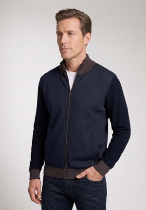 Mann mit kurzen braunen Haaren, der eine marineblaue Reißverschlussjacke mit grauem Kragen und Bündchen sowie dunkle Jeans trägt und vor einem schlichten Hintergrund steht.