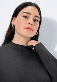 Junge Frau mit langen dunklen Haaren trägt silberne Creolen und einen dunklen Hochkragenpullover und berührt mit ihrer Hand den Kragen.