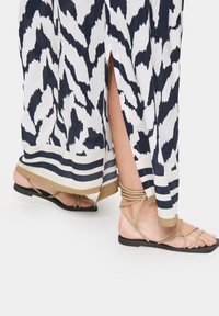 Maxi-jurk met een wit en marineblauw abstract patroon, zijsplitten en een blauwe gestreepte zoom; gecombineerd met neutrale sandalen met banden.