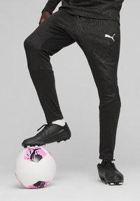 Zwarte voetbalbroek met een gestructureerd patroon, gecombineerd met zwarte voetbalschoenen, één voet op een witte bal met roze en zwarte accenten.