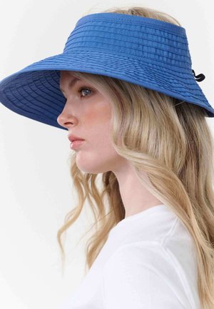 Woman with long blonde hair wearing a wide-brimmed blue sun hat and a white top, facing left against a plain background.