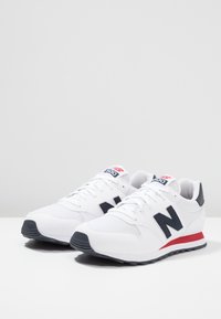 Un paio di sneakers bianche New Balance 500 con logo "N" nero, striscia rossa sulla suola e accenti neri sul tallone e sulla punta, su una superficie bianca.