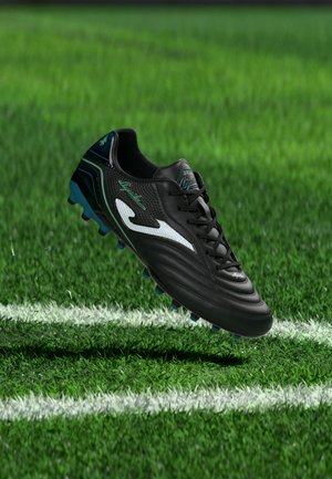 Schwarzer Fußballschuh mit weißem Logo und blauen Stollen, der über grünem Gras in der Nähe weißer Spielfeldlinien schwebt.