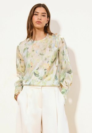 Frau trägt eine transparente, langärmelige Bluse mit Blumenmuster und Puffärmeln, kombiniert mit hoch taillierten, cremefarbenen Hosen mit weitem Bein.