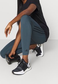 Czarna koszulka, turkusowe legginsy z trzema czarnymi paskami, czarno-białe buty sportowe i czarna hantelka w ręku.