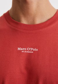 Red cotton t-shirt featuring a crew neck and short sleeves. White text reads "Marc O'Polo EST. IN STOCKHOLM" on the chest.