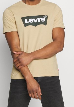 Person trägt ein beiges Levi's T-Shirt und dunkle Hosen, steht mit verschränkten Armen vor einem schlichten hellen Hintergrund.