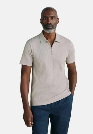 Man with gray beard wearing light beige short-sleeve zip collar shirt and dark blue pants standing against white background.