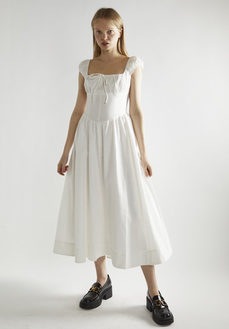 Witte, knie-lange jurk met een vierkante halslijn en pofmouwen, met een geraffineerde taille en vloeiende rok. Gedragen met chunky zwarte loafers.
