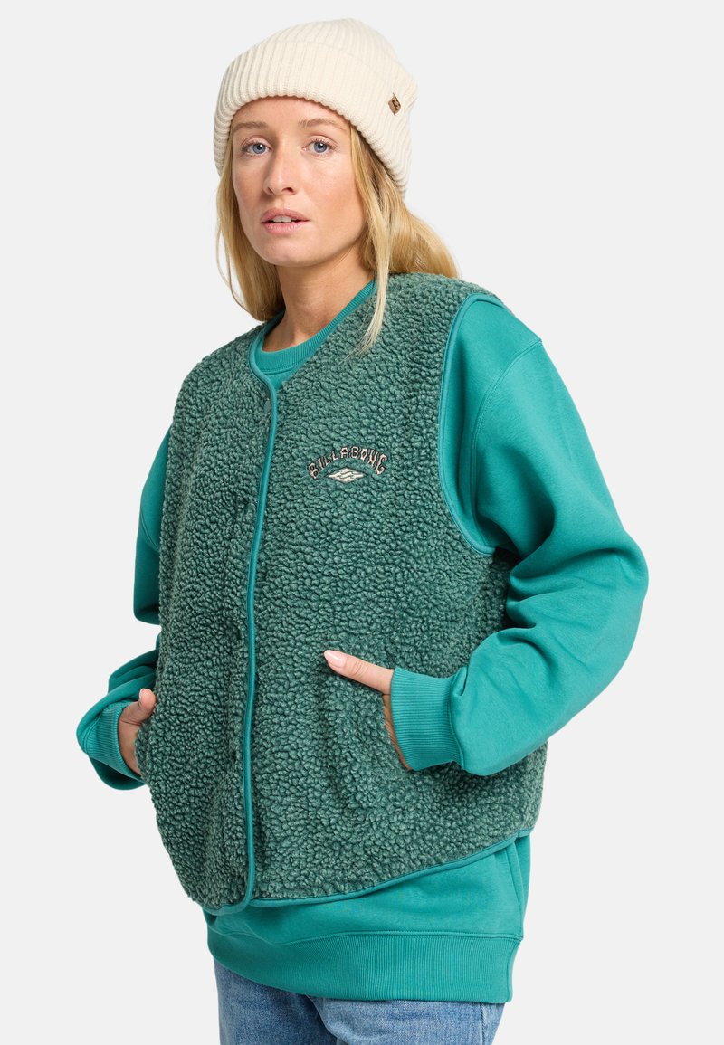 Teal fleece vest med en teksturert overflate, kontrasterende ribbestrikkede mansjetter og logo-detalj. Brukt over en teal sweatshirt med sidelommer.