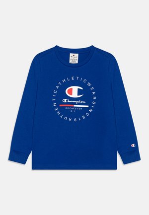 Camiseta de manga larga en algodón azul con un logo circular de Champion en blanco y rojo. Presenta un cuello redondo y puños acanalados.