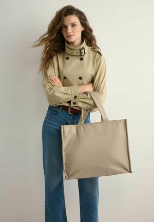 Kobieta z długimi falującymi włosami, ubrana w beżową, dwurzędową marynarkę i niebieskie dżinsy, trzymająca dużą beżową torbę typu tote z dwoma uchwytami.