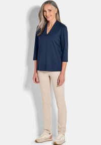Navyblaue 3/4-Ärmelbluse mit V-Ausschnitt, kombiniert mit hellbeigen, figurbetonten Hosen und beigen Sneakers mit strukturierten Sohlen.