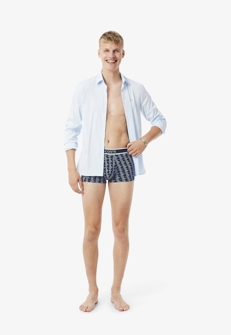 Modelo vistiendo una camisa de botones azul claro, abierta sobre unos pantalones cortos de boxeo con patrón en color azul marino y cinturilla negra; de pie descalzo, sonriendo.