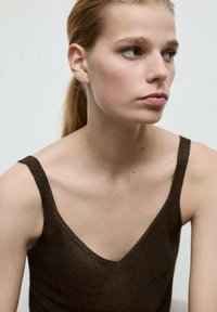 Maiou fără mâneci, maro închis, cu design decoltat în V. Textură moale tricotată, cu un luciu subtil. Model cu păr drept, pozând pe un fundal deschis.