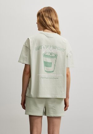 Hosszú hajú nő világoszöld rövidnadrágban és bő, túlságosan nagy pólóban áll, amelyen jeges matcha ital grafikája és szövege látható a hátulján, háttal a néző felé.