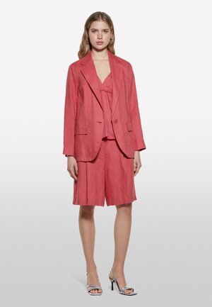 Blazer en lin rose et short assorti, avec une coupe décontractée. Le blazer possède des épaules padding et deux poches avant. Sandales à talons hauts argentées.