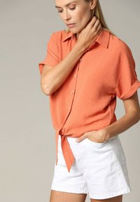 Oranje kortef-blouse met knopen en een gestrikte voorkant, gecombineerd met witte geborduurde shorts met subtiele patronen en zijzakken.