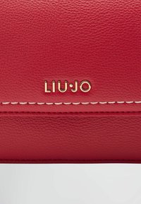 Cartera de cuero texturizado rojo con un logotipo en tono dorado "LIU·JO" y costuras blancas a lo largo del borde. Diseño compacto con una superficie lisa.