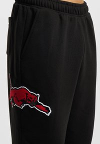 Svarta sweatpants med elastiskt midjeband, med en stor röd broderad logotyp av ett hoppande djur på vänster lår. Slät textur.