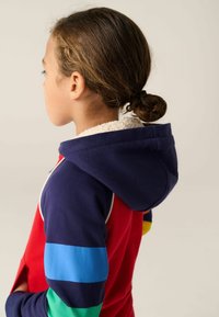 Felpa colorata con corpo rosso, cappuccio blu navy e maniche a righe blu, verdi e gialle. Fodera interna testurizzata, coda di cavallo scura.