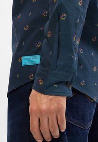 Ναυτικό μπλε πουκάμισο με μικρά, πολύχρωμα λουλουδάτα σχέδια. Διαθέτει κουμπωτό μανσέτα και επώ νυμη ετικέτα σε τυρκουάζ. Υφασμάτινο υλικό από βαμβάκι.