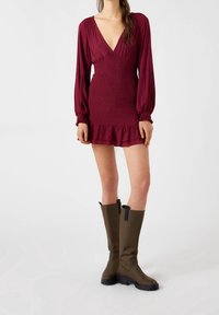 Vestido burdeos de manga larga con escote en V profundo y cuerpo fruncido, con un dobladillo de volantes en varias capas; acompañado de botas de caña alta color verde oliva.