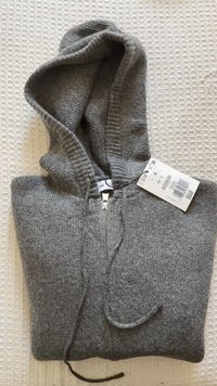 Siva pletena jopa s sprednjo zadrgo, vrvico in priloženo značko, postavljena na teksturirano površino. Ima kapuco in rebraste manšete.