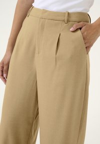 Beige wijde broek met een geplooide voorkant, een op maat gemaakte taille met riemlussen en zijzakken, gemaakt van een gladde stof.
