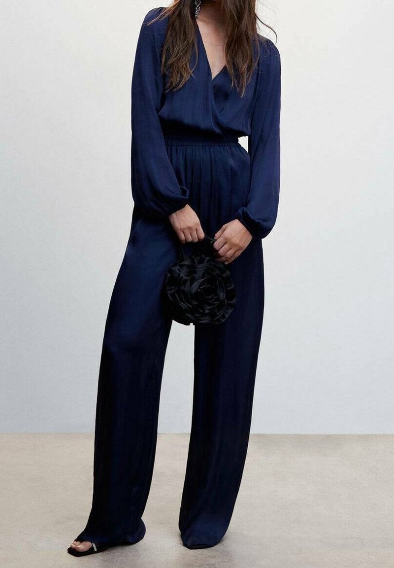 Femme portant une combinaison bleu marine à manches longues tenant un petit sac à main rond noir, debout sur un sol beige contre un mur blanc.