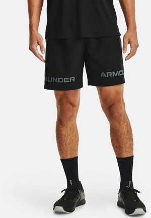 Męski model nosi czarne krótkie spodenki Under Armour, czarne skarpety za kostkę i czarne sportowe buty, stoi z lekko rozstawionymi nogami.