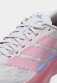 Sneaker blanche avec des rayures roses, logo bleu sur la languette, lacets blancs et semelle rose avec accent bleu, présentée sur un fond blanc.