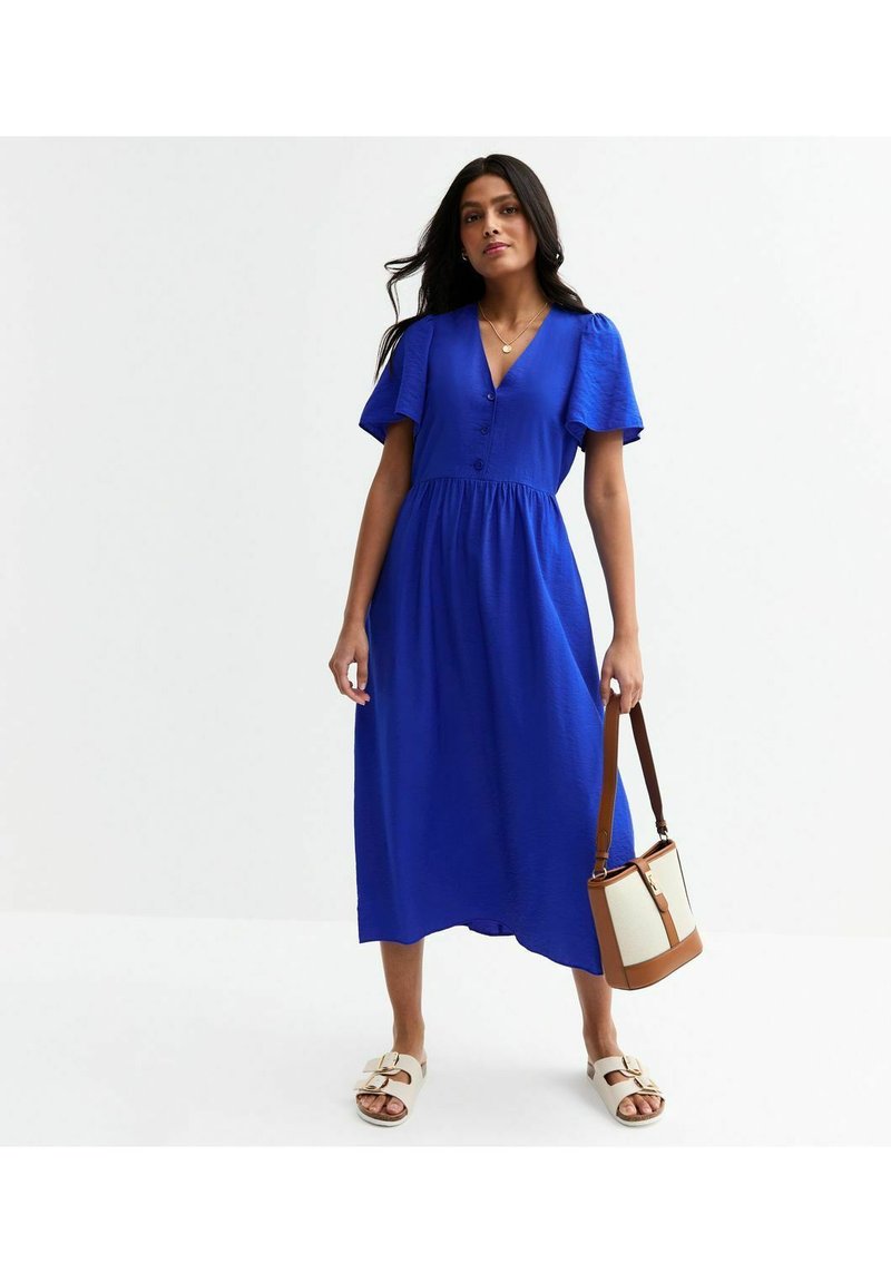 Vestido midi azul con mangas cortas tipo volantes, detalles de botones y una cintura fruncida. Combinado con sandalias blancas y un bolso cruzado marrón.