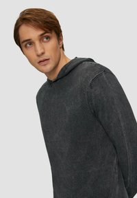 Haut à capuche gris en tissu texturé, avec une capuche, des poignets côtelés et une coupe surdimensionnée. Des variations subtiles de couleur ajoutent de la profondeur.