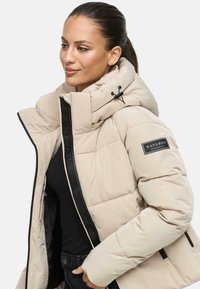 Femme portant une doudoune beige avec un col montant, une fermeture éclair noire et un écusson Navahoo sur la manche, posant avec les mains dans les poches.