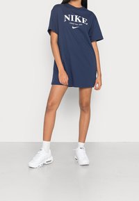 Námořnické modré oversized tričko Nike s bílým logem; krátké rukávy a kulatý výstřih; nošeno s bílými teniskami a ponožkami.