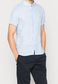 Lichtblauw linnen overhemd met korte mouwen, een button-down kraag en een klein logodetail. Draagt donkere broek, handen in de zakken.