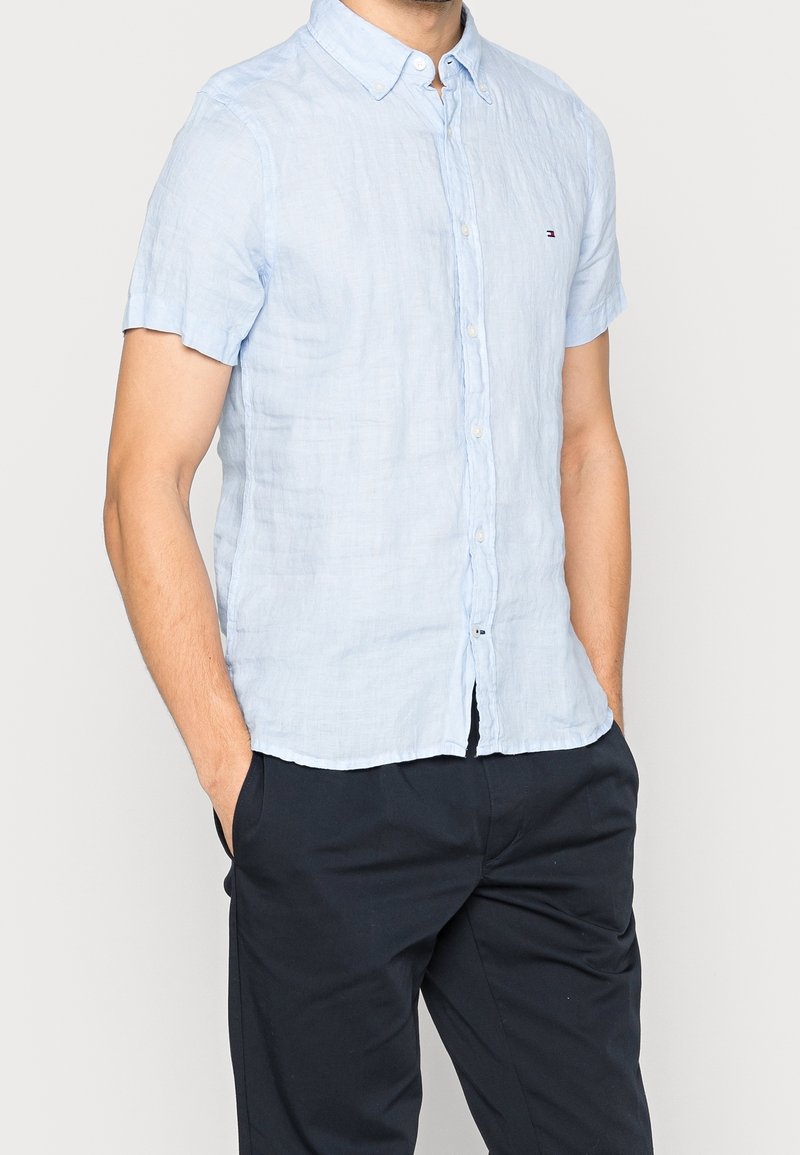 Lichtblauw linnen overhemd met korte mouwen, een button-down kraag en een klein logodetail. Draagt donkere broek, handen in de zakken.