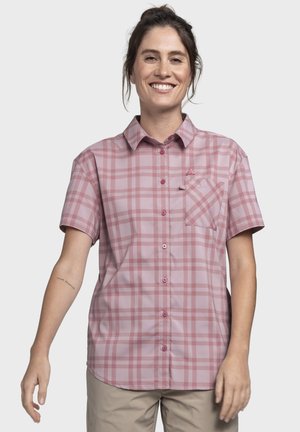 Frau lächelnd, trägt ein kurzärmeliges, rosa kariertes Hemd mit Knöpfen und beige Hosen, steht vor einem schlichten hellen Hintergrund.