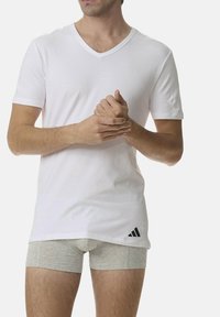 Witte V-hals T-shirt met korte mouwen, gemaakt van lichtgewicht materiaal. Heeft een zwart Adidas-logo op de linkeronderkant. Grijze onderbroek is zichtbaar.